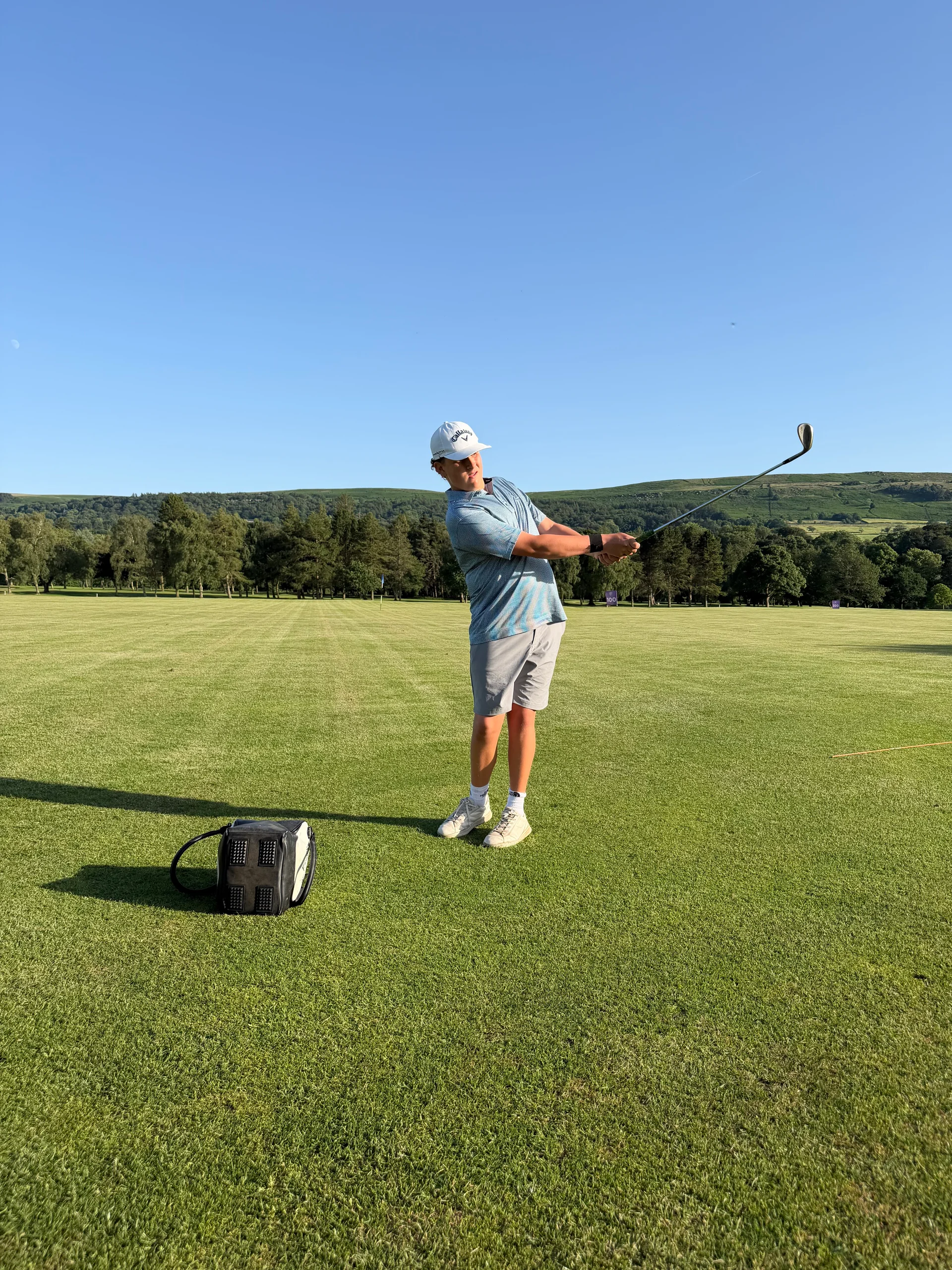 A man swings a golf club on a green field with trees and hills in the background.