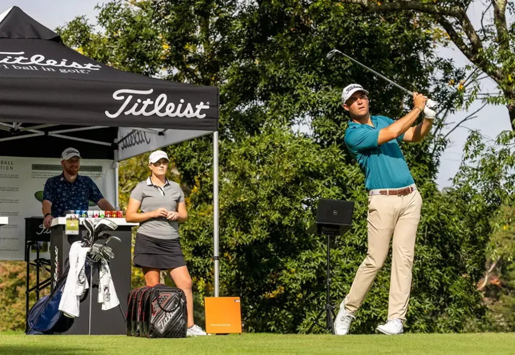 A golfer swings during a golf event with a Titleist tent, a woman and a man standing nearby on a grassy field.