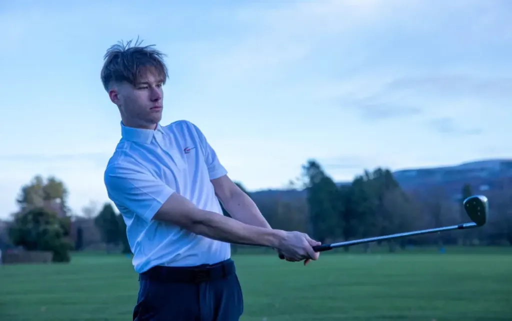 A young man swings a golf club on a green field with hills in the background.