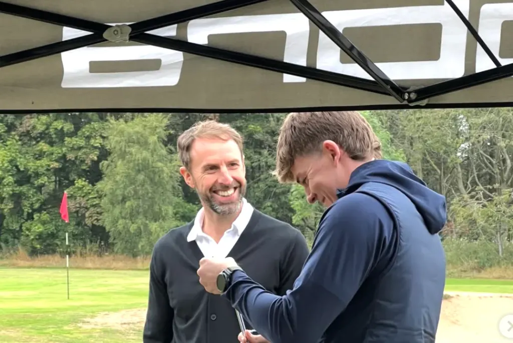 Two men are talking and looking at a device, standing on a green field with a golf flag in the background.